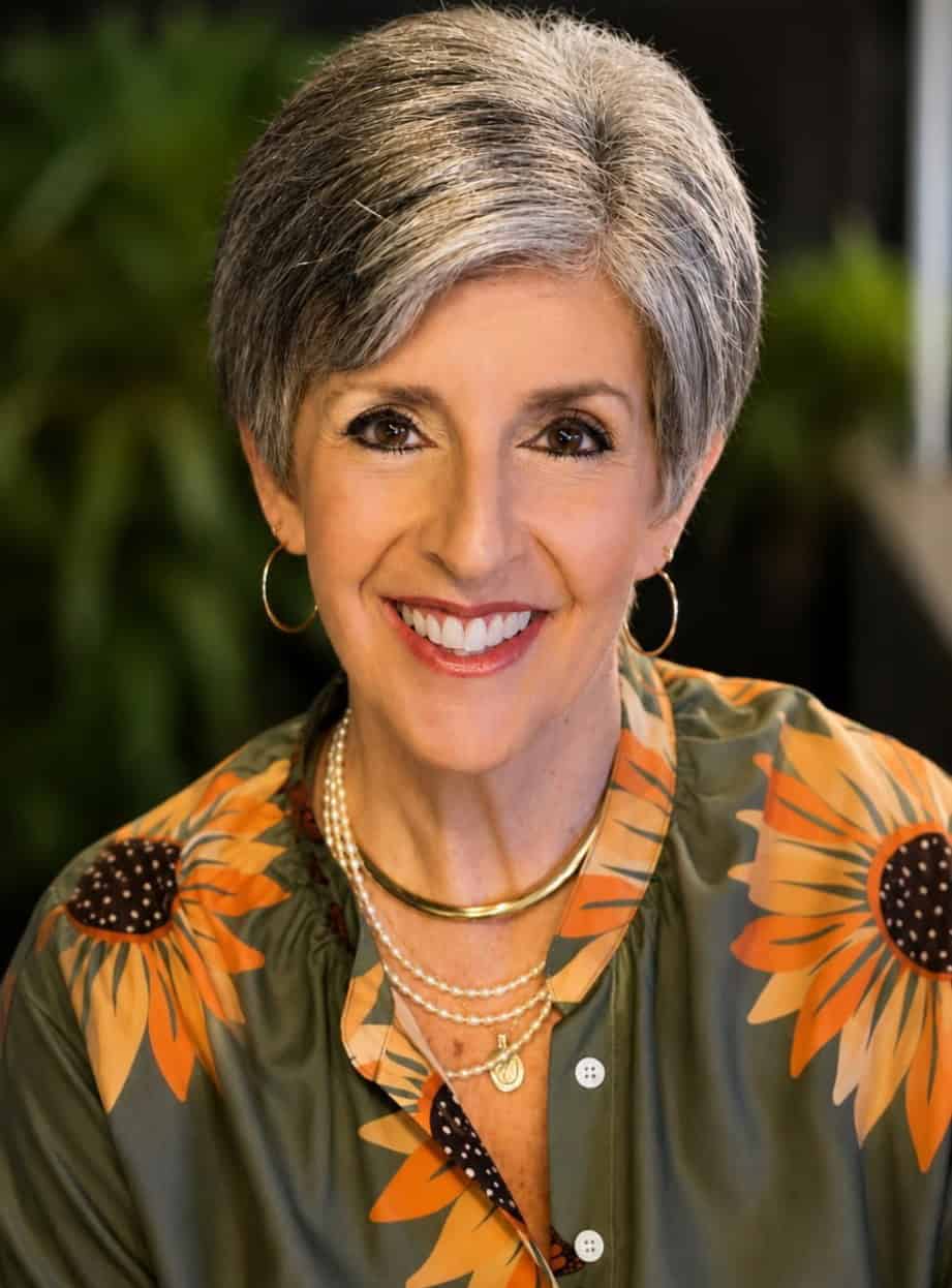 Professional portrait of Wanda Roussel, smiling woman with short gray hair, wearing a floral blouse and jewelry, in a well-lit indoor setting.